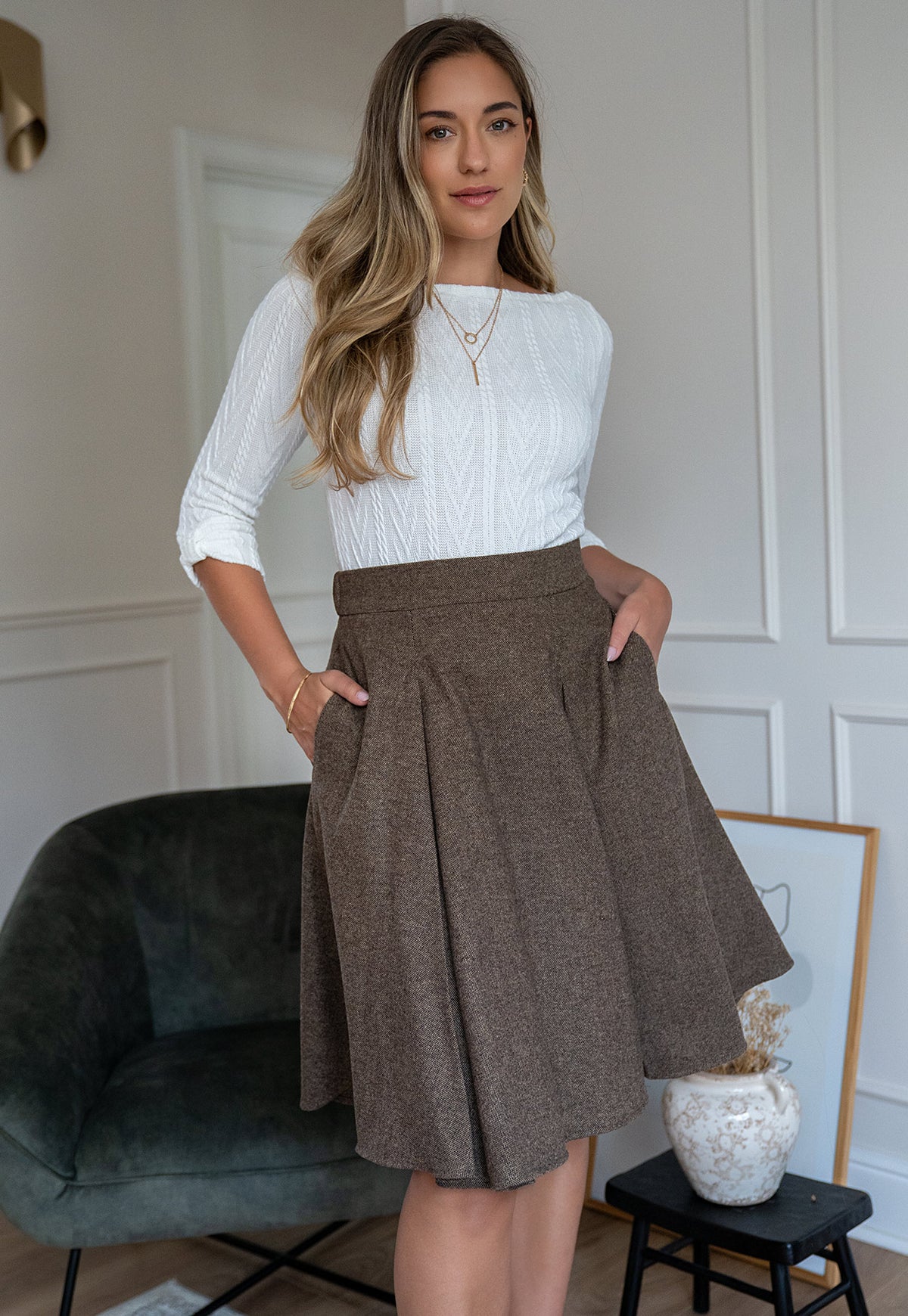 A woman stands indoors wearing a Women’s Aniz ivory textured cable-knit top with boat neckline and three-quarter sleeves by Elizabeth Lozano. and a brown pleated high-waisted skirt with side pockets. The setting includes a dark green velvet chair, a small black table with a white vase, and a white paneled wall.